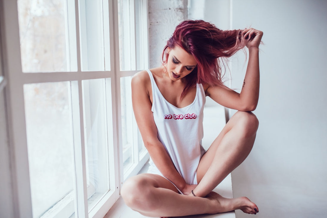Woman with red hair sitting on a windowsill, wearing a 'no bra club' tank top in white with red font.